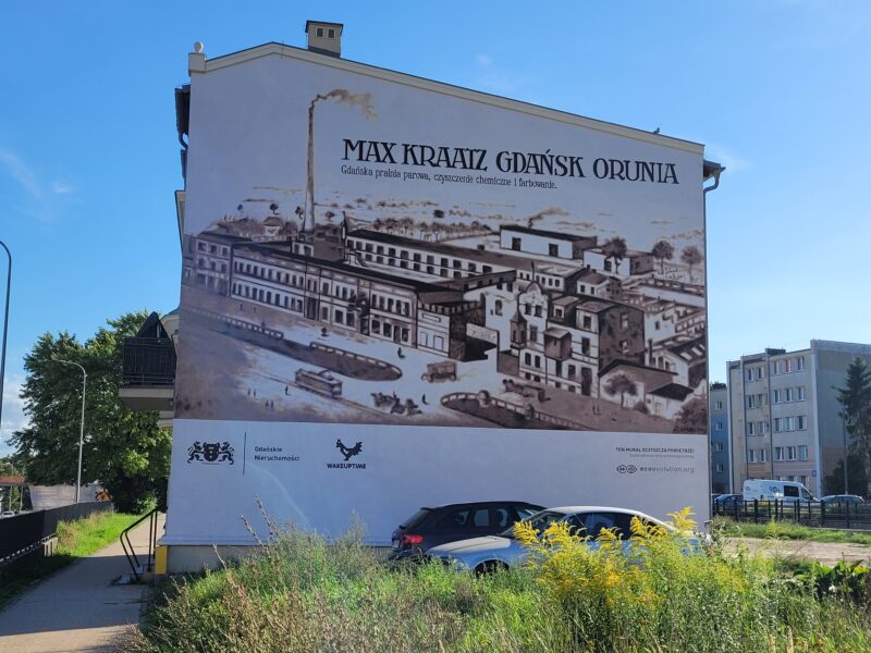 Mural na ścianie budynku przedstawiający namalowaną na brązowo fabrykę z wysokim kominem.