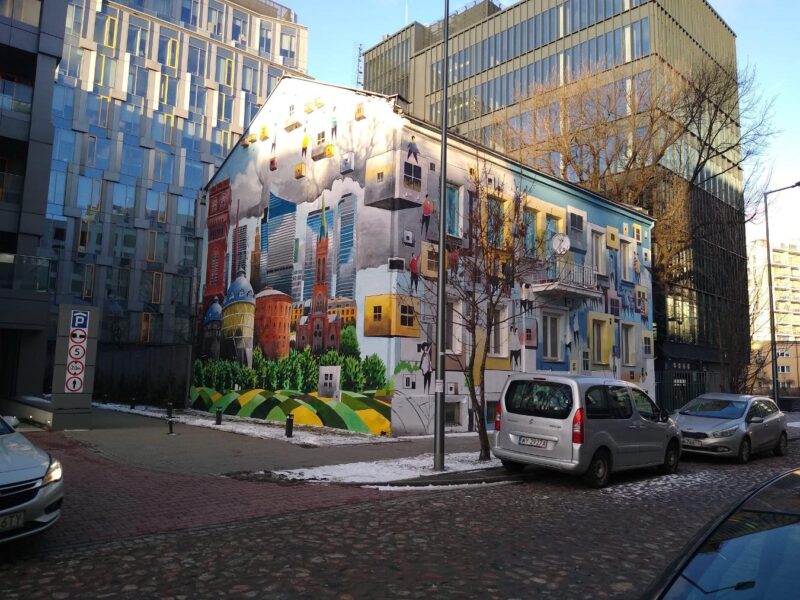 Budynek piętrowy pokryty muralem. Przed budynkiem stoją dwa samochody osobowe.
